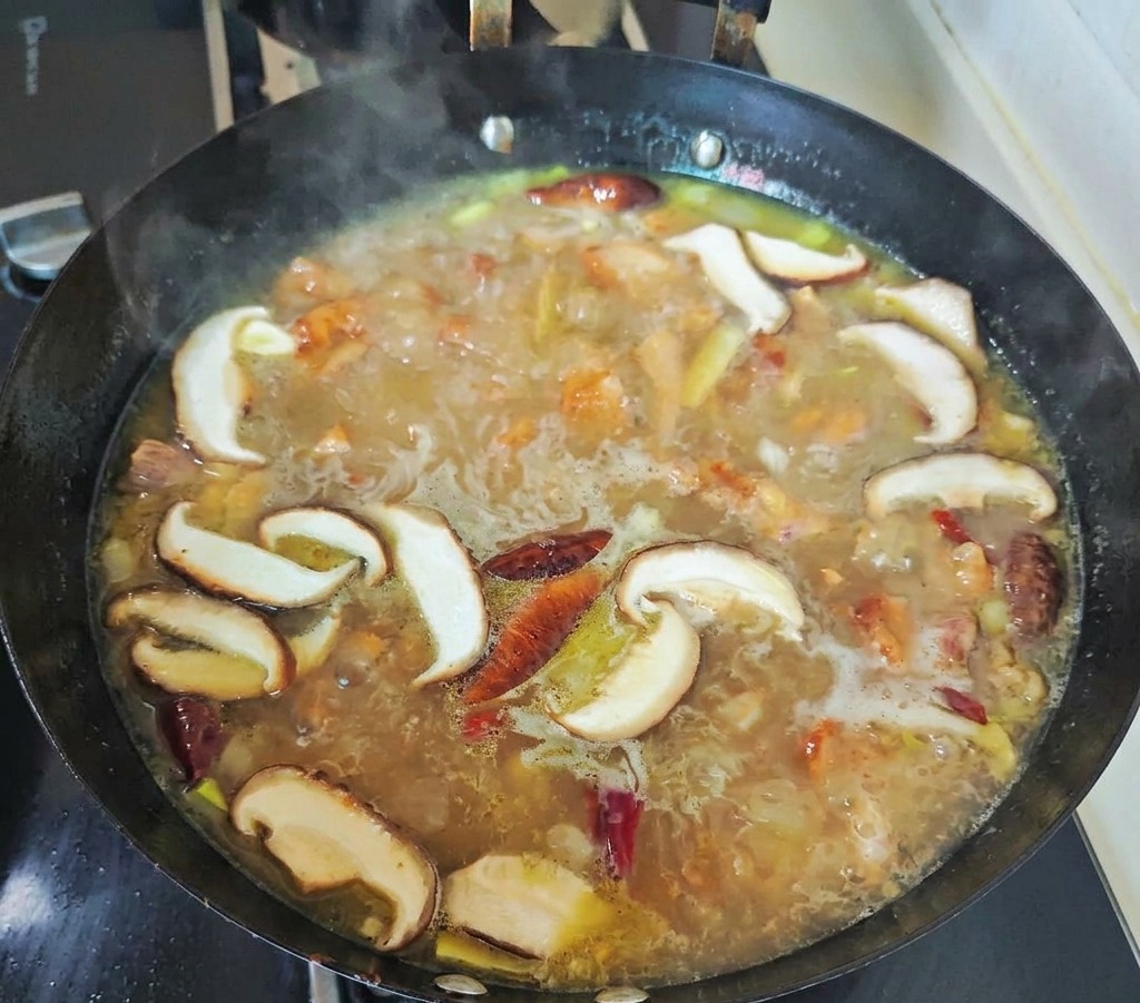 Sliced shiitake mushrooms and red chilies simmering in a bubbling golden chicken broth within a black wok.