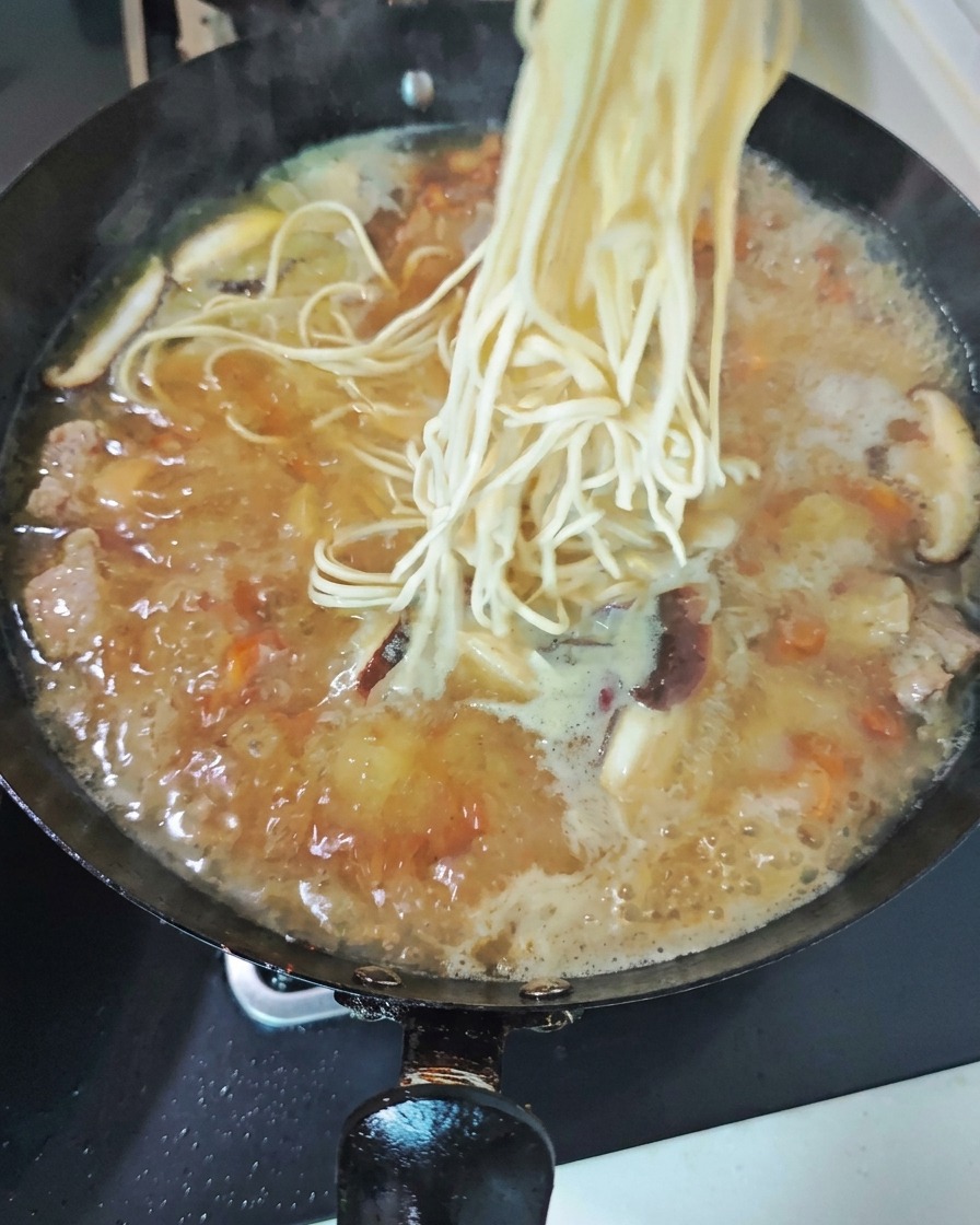A bundle of fresh, pale noodles being dropped into a bubbling wok of savory chicken soup filled with sliced mushrooms.