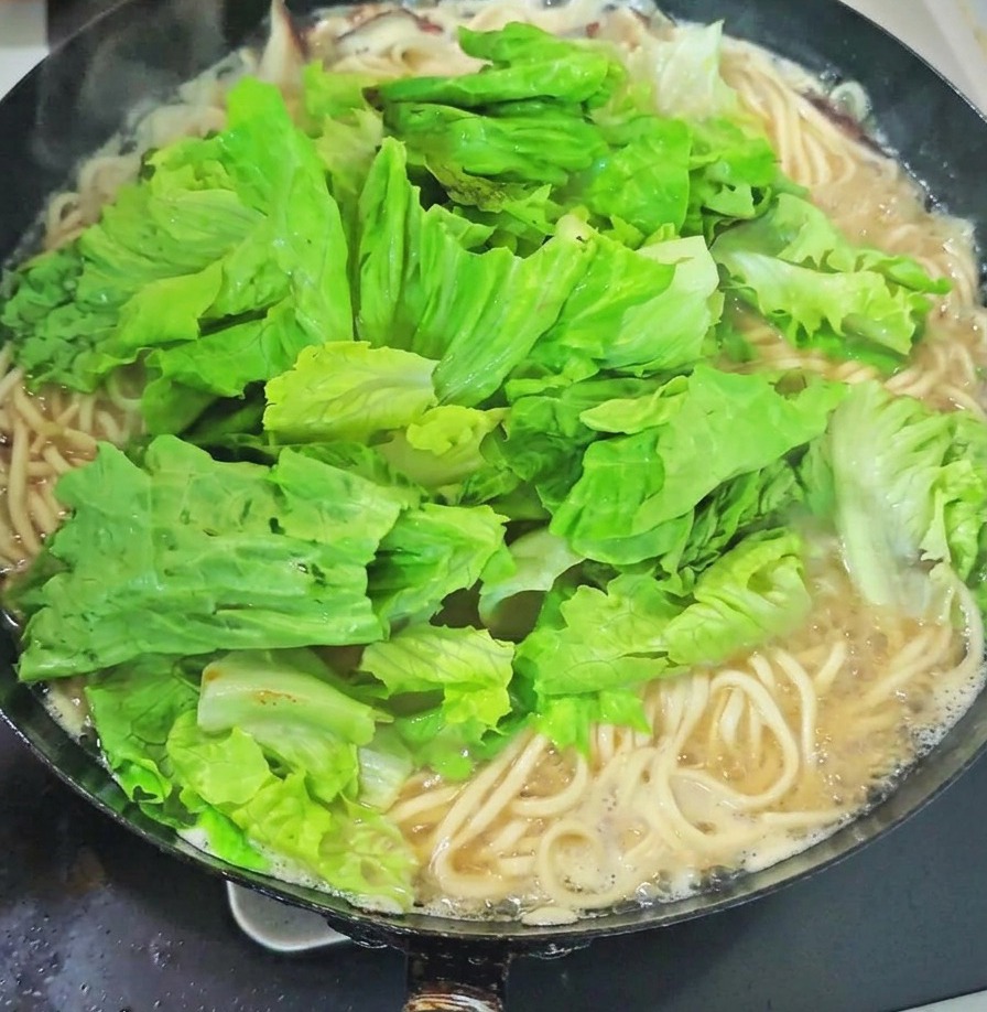 A large pile of fresh green leafy vegetables and garlic sprouts sitting on top of noodles in a boiling wok of soup.