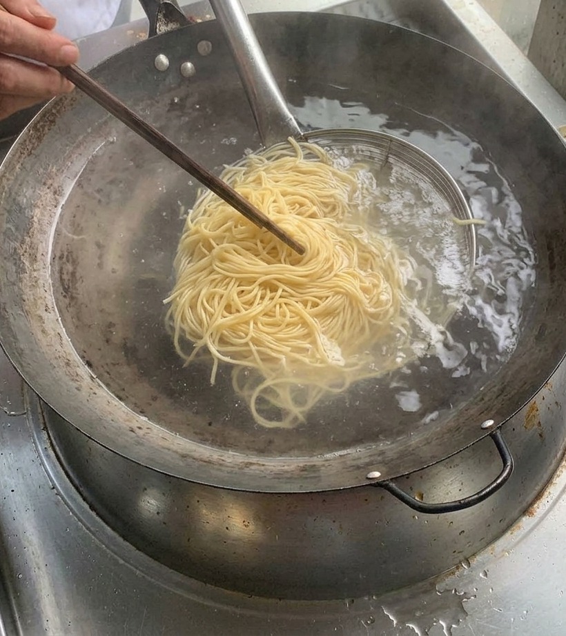 Cooked noodles held in a metal strainer over a pot of boiling water.