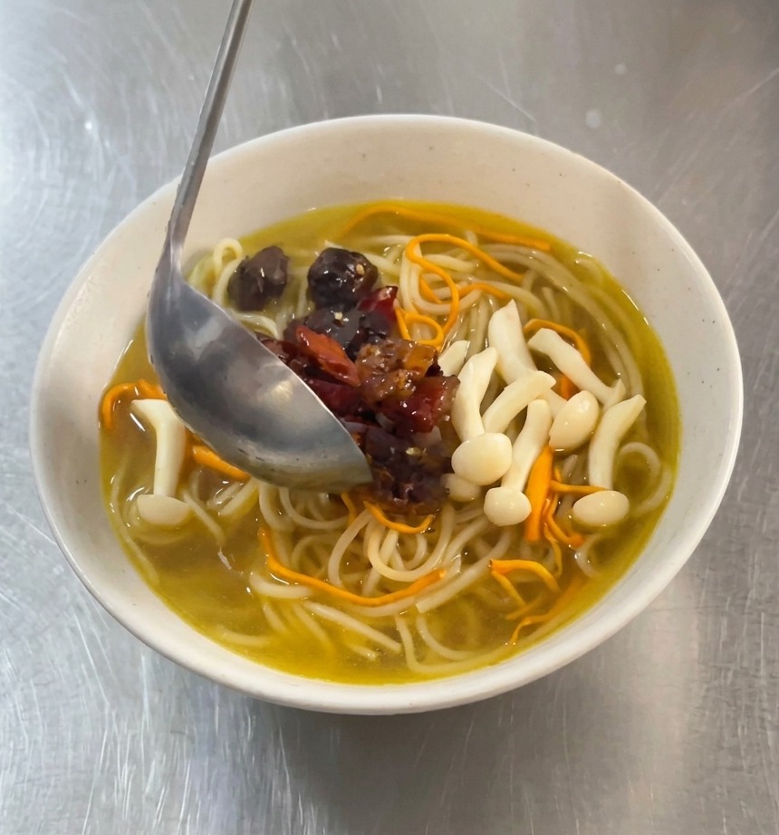 A metal ladle pouring pieces of red-marinated spicy chicken and dried chilies onto a bowl of white noodles in a golden mushroom broth.