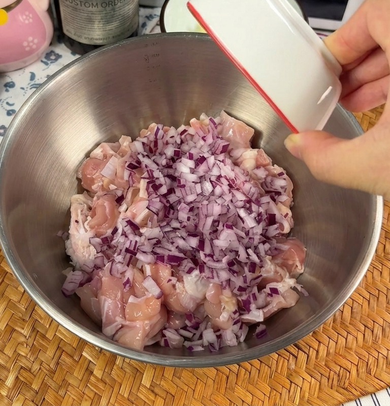 A hand pouring a small dish of chopped red onions and minced garlic over raw chicken chunks in a metal mixing bowl.