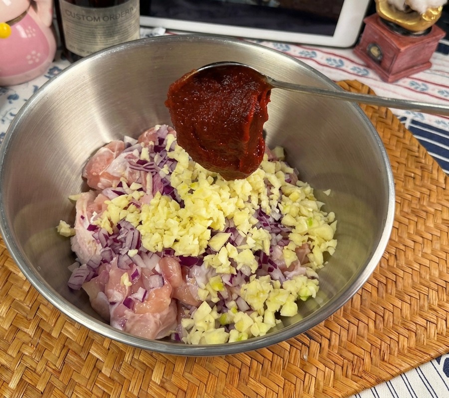 A metal spoon holding a large dollop of vibrant red Gochujang paste over a bowl containing raw chicken, garlic, and onions.