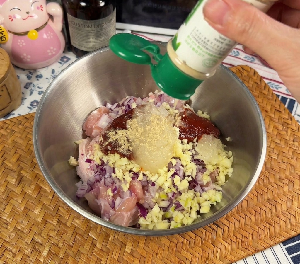 A person sprinkling white pepper powder from a green shaker into a metal bowl containing raw chicken and chopped red onions.