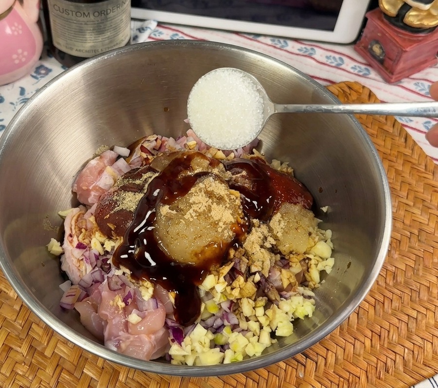 A spoonful of granulated white sugar being added to a bowl of chicken marinating in a thick red sauce.