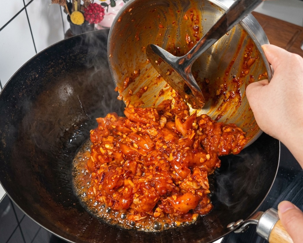 Marinated chicken chunks in a bright red sauce being poured from a metal bowl into a hot steaming black wok.