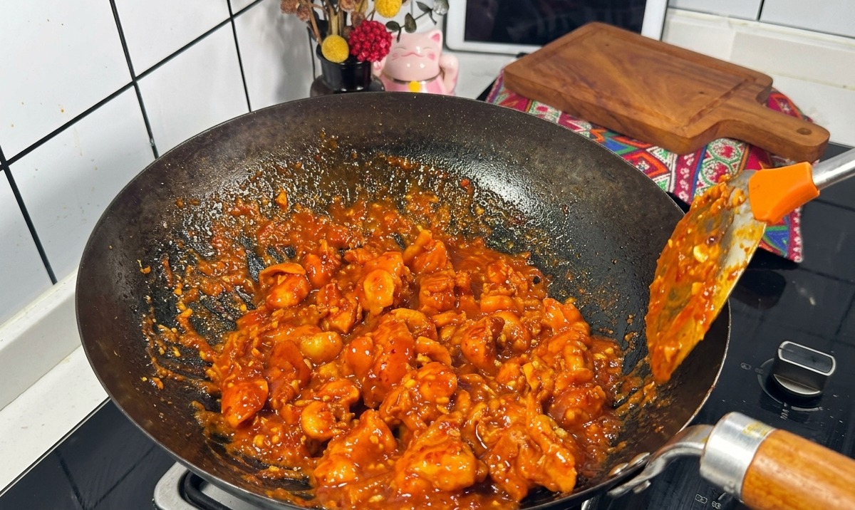 Chicken pieces coated in a thick, bubbling red sauce being stir-fried in a black wok with a metal spatula.