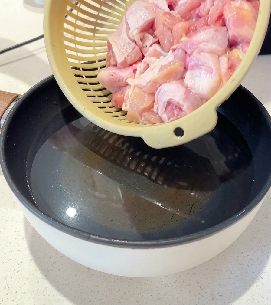A person pouring raw chicken pieces from a colander into a pot of water containing sliced ginger and scallions.