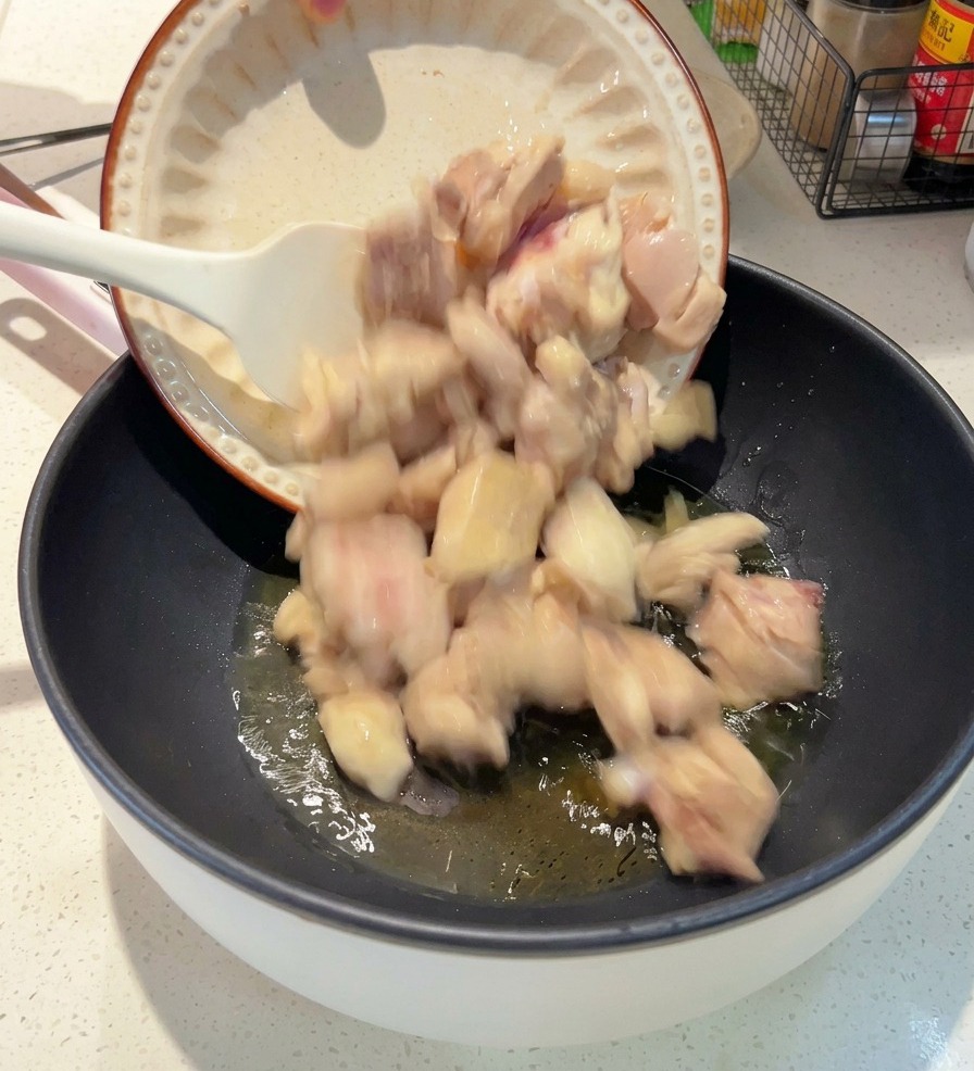 A white plate pouring blanched chicken pieces into a dark pan with shimmering oil.