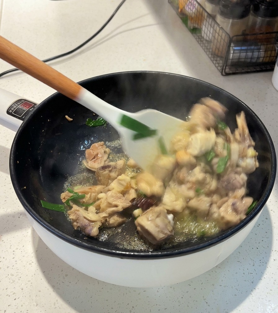 A white spatula stirring chicken pieces, scallions, and star anise in a pan over heat.