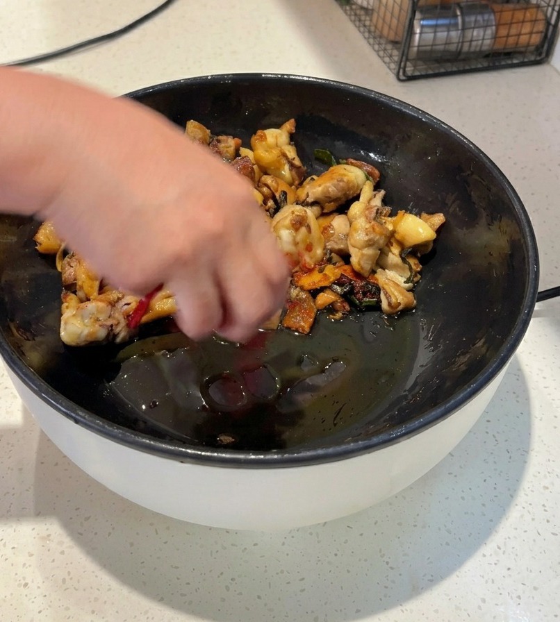 A person stir-frying chicken pieces in a black pan with whole dried red chilies and bright red chili powder.