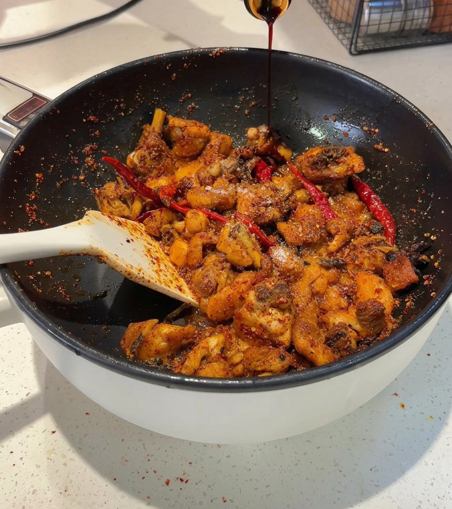 Dark soy sauce being poured from a bottle into a frying pan containing spicy chicken pieces.