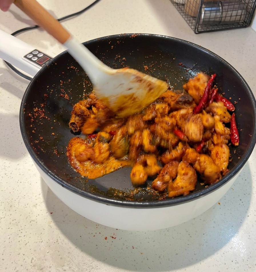 A white spatula tossing pieces of chicken in a dark red sauce inside a pan until every piece is coated.
