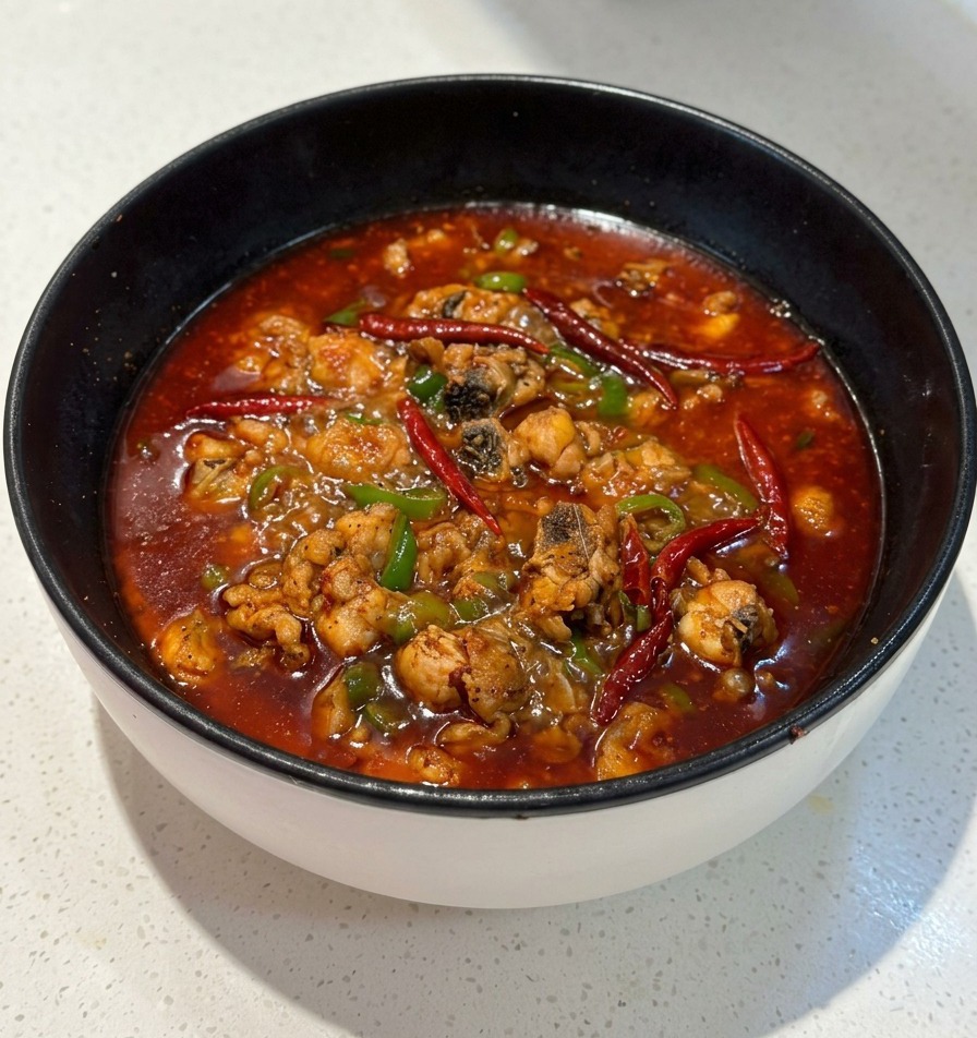 A black pan filled with a bubbling, spicy red broth containing chicken pieces and green chili segments.
