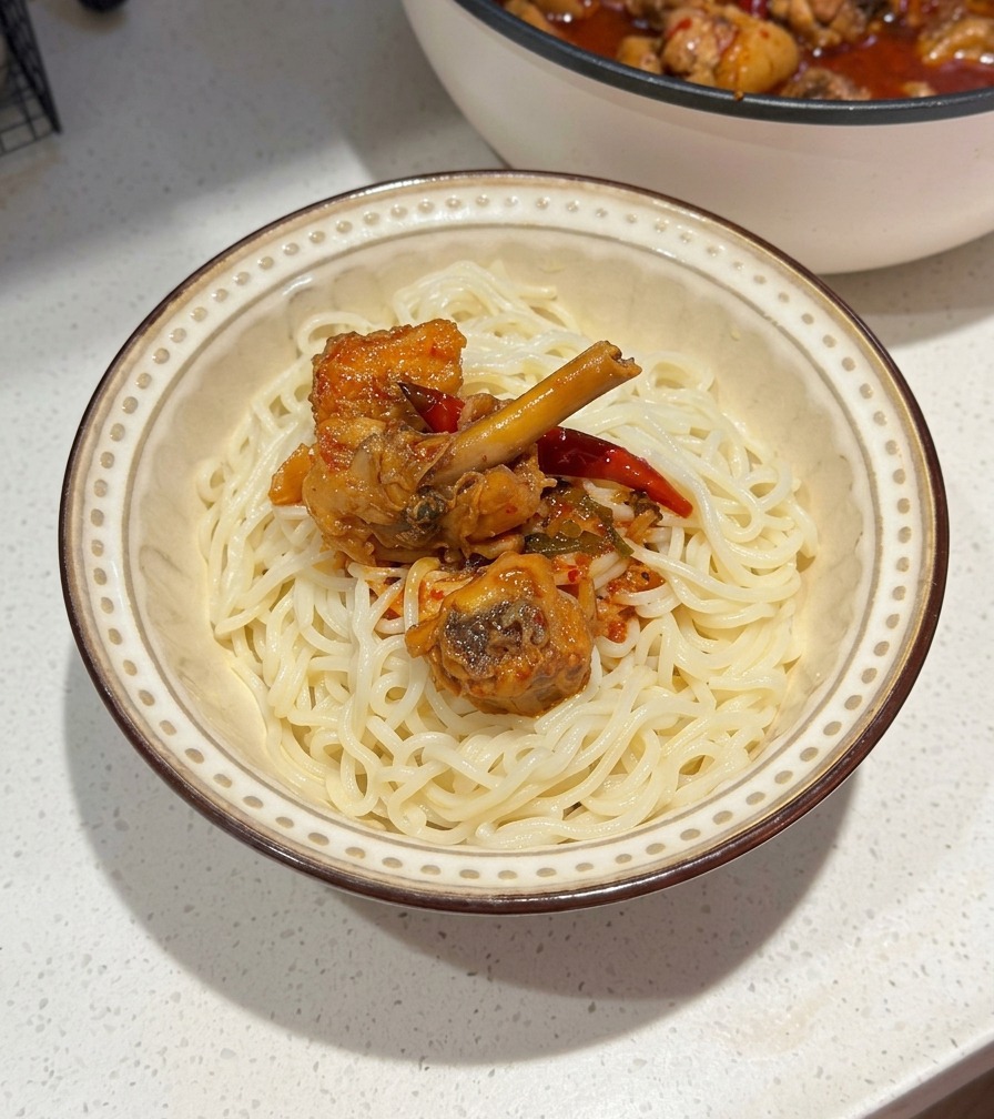 A ceramic bowl of white noodles topped with spicy chicken and red chili sauce, with the cooking pan visible in the background.