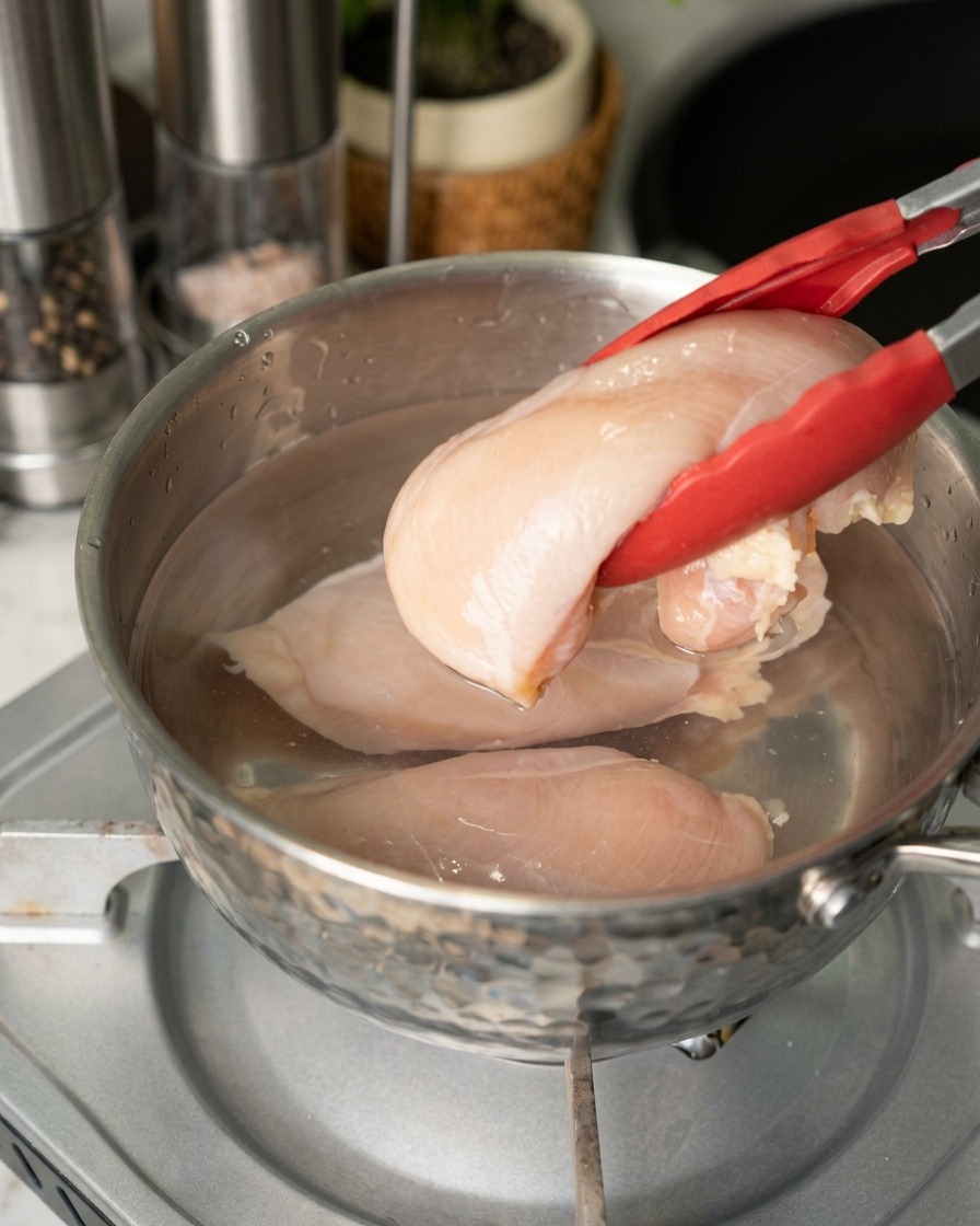 A person using red-tipped tongs to place raw, pale pink chicken breasts into a stainless steel pot of water.