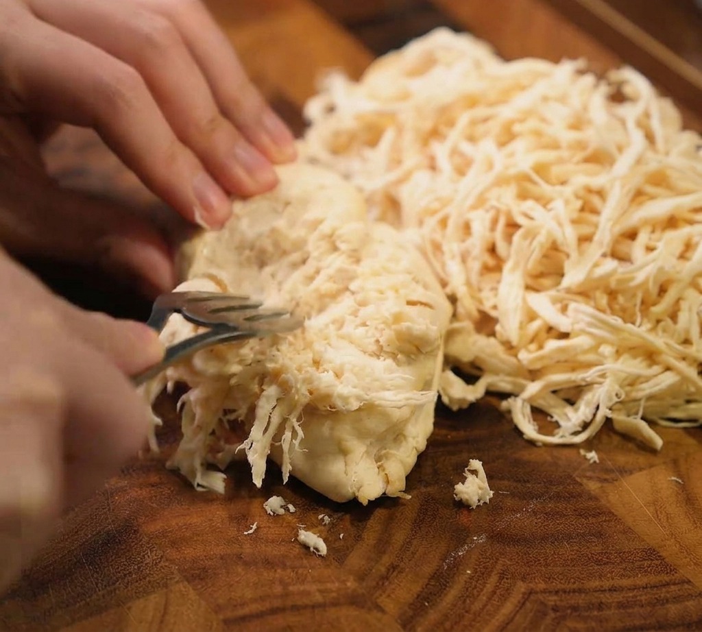 A pair of metal forks being used to shred a whole cooked chicken breast into thin strips on a dark wood cutting board.