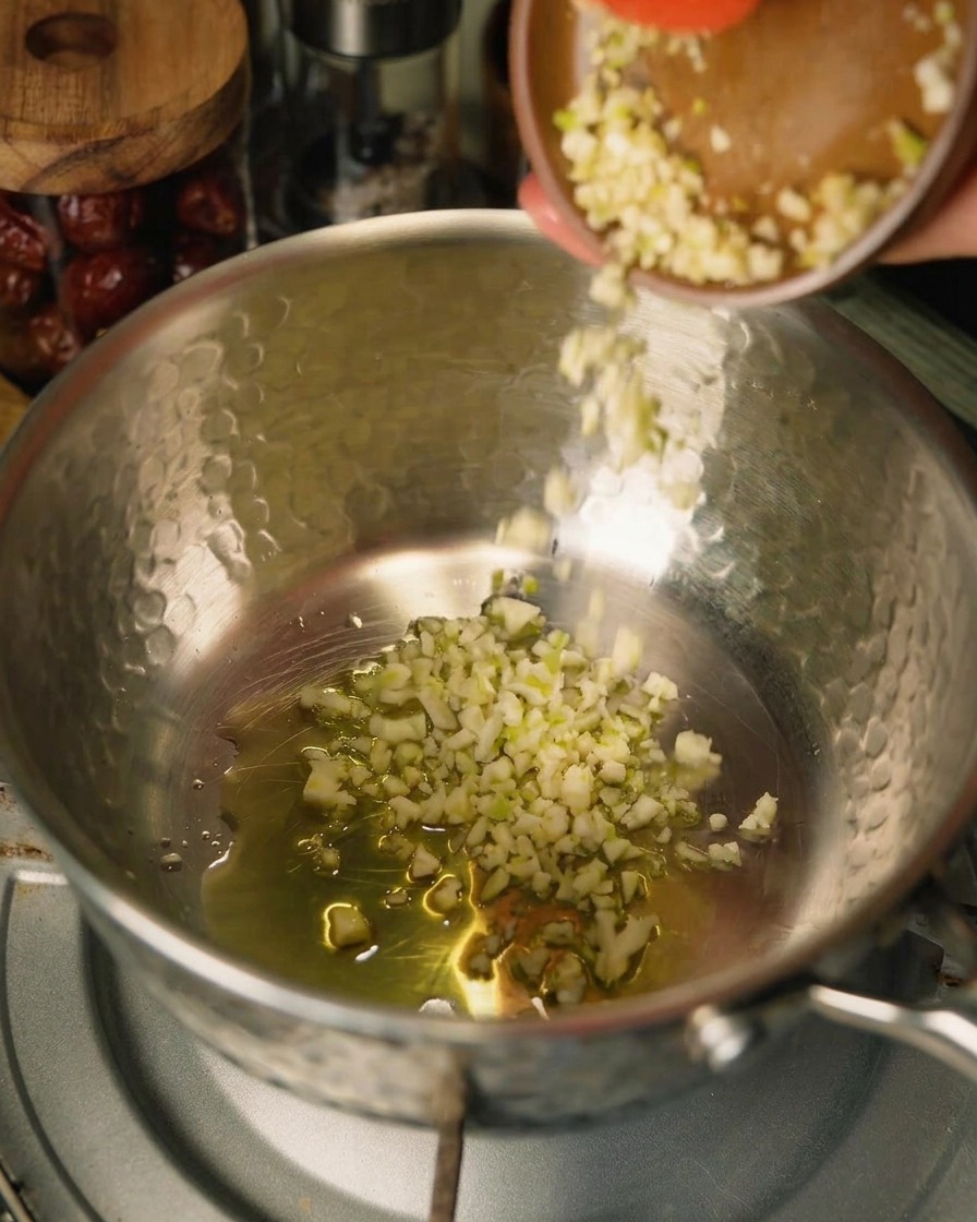 Minced garlic being poured from a small brown bowl into a pot with oil and onions, with a red spatula edge visible.