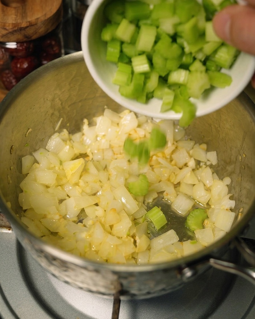 Diced celery being added from a white bowl into a pot that already contains sautéed onions and bright orange shredded carrots.