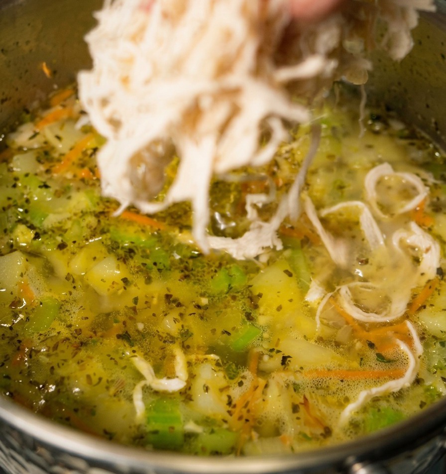 A hand dropping shredded white chicken meat into a pot of simmering vegetable soup with visible carrots and celery.