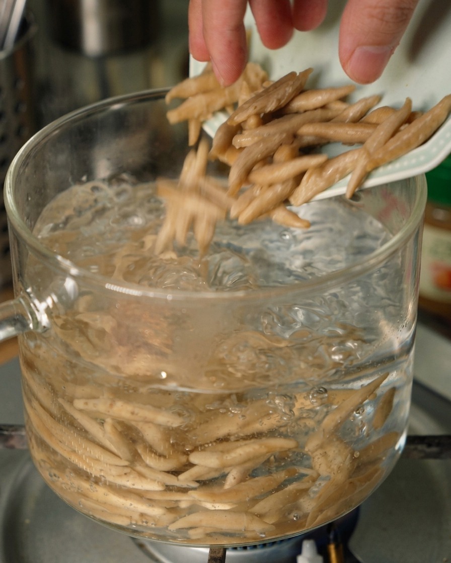 Tapered oat noodles falling from a small plate into a clear glass pot of bubbling boiling water.