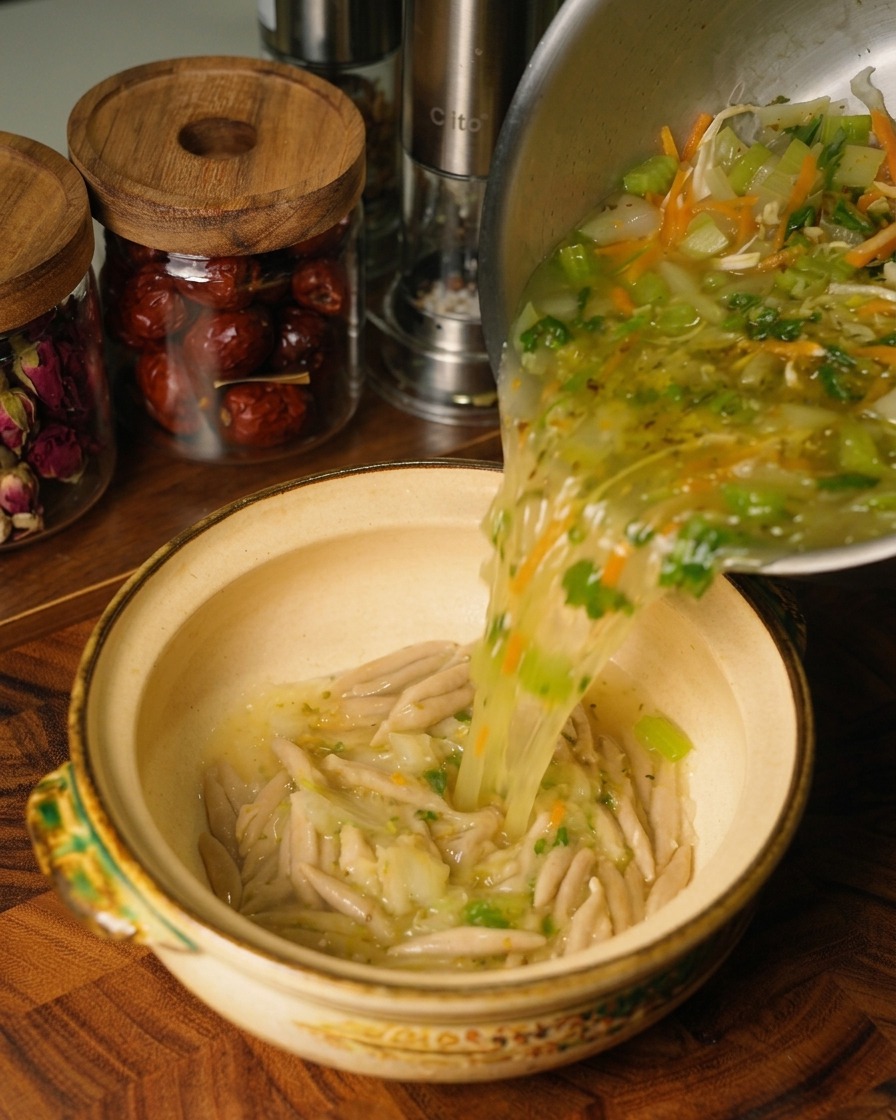 Hot chicken and vegetable soup being poured from a stainless steel pot into a ceramic bowl containing cooked oat noodles.