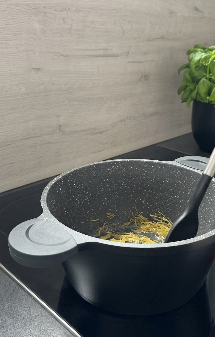 Small needle-shaped pasta being stirred with a black spatula in a grey granite-patterned pot as it toasts to a golden brown.