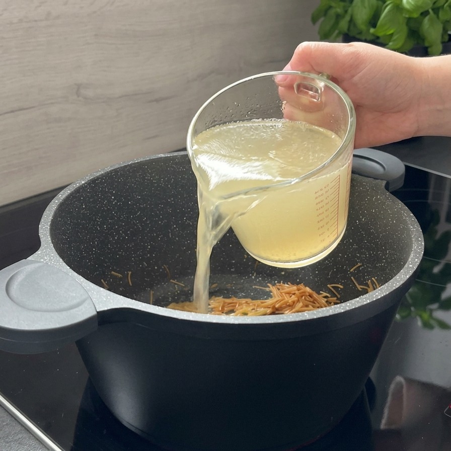 Clear chicken broth being poured from a glass measuring cup into a black pot containing toasted brown pasta.