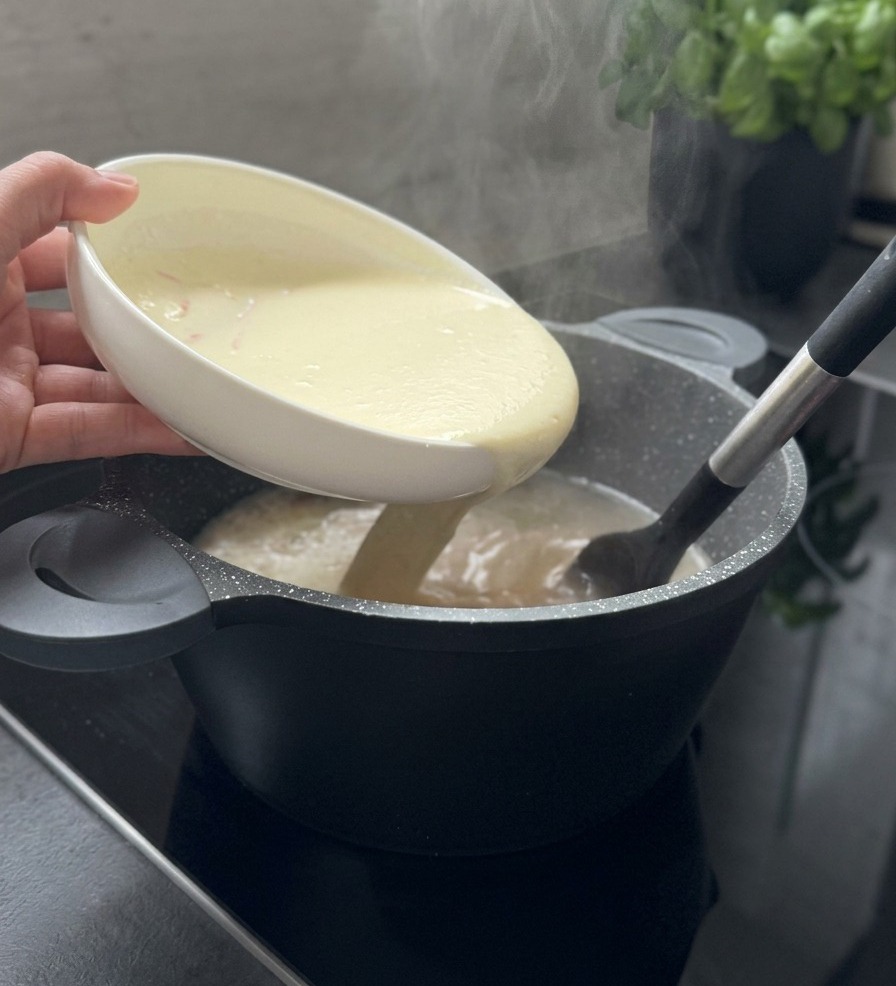 A creamy white thickening mixture being poured from a white bowl into a steaming pot of soup while being stirred with a black spatula.