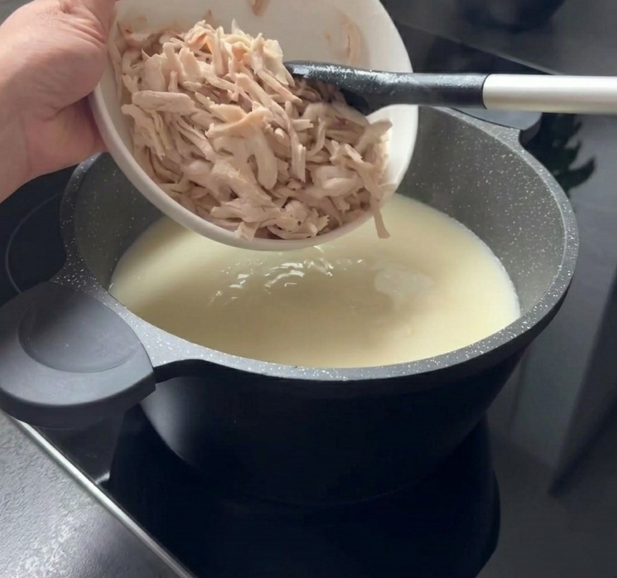 Shredded pieces of cooked chicken being added from a white bowl into a pot of creamy white soup.