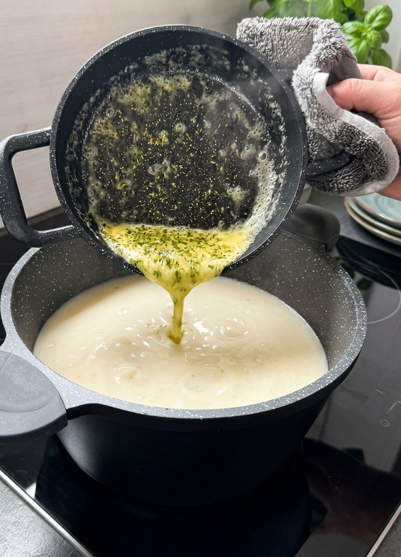Melted yellow butter with green dried mint flakes being poured from a small black pot into a creamy white soup.