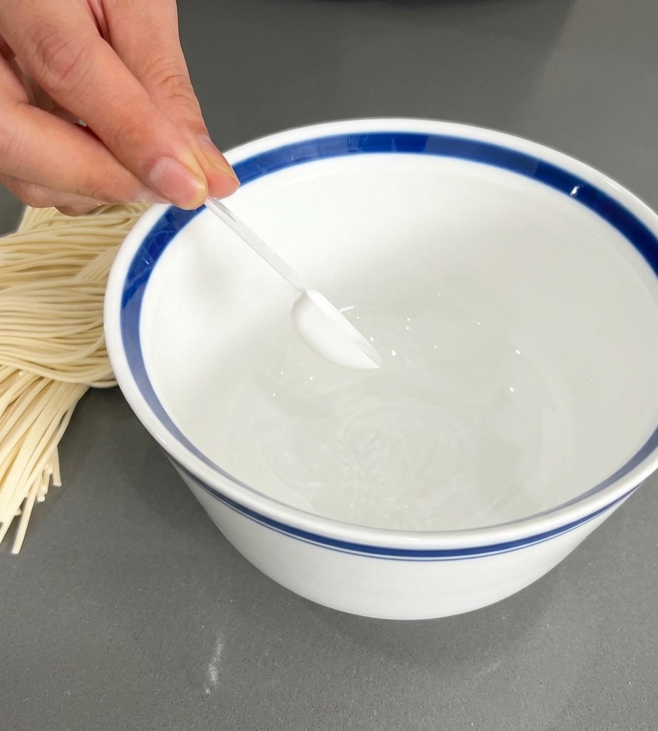 A hand holding a small clear spoon, placing white seasoning powder into a white bowl with a distinct blue rim.