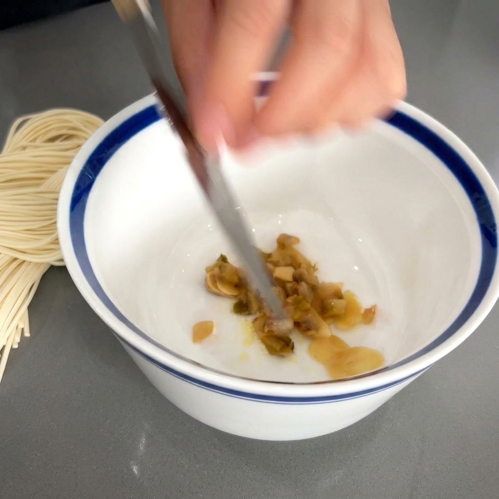 A knife blade sliding chopped brown pickled vegetables into a white bowl containing garlic and seasoning.