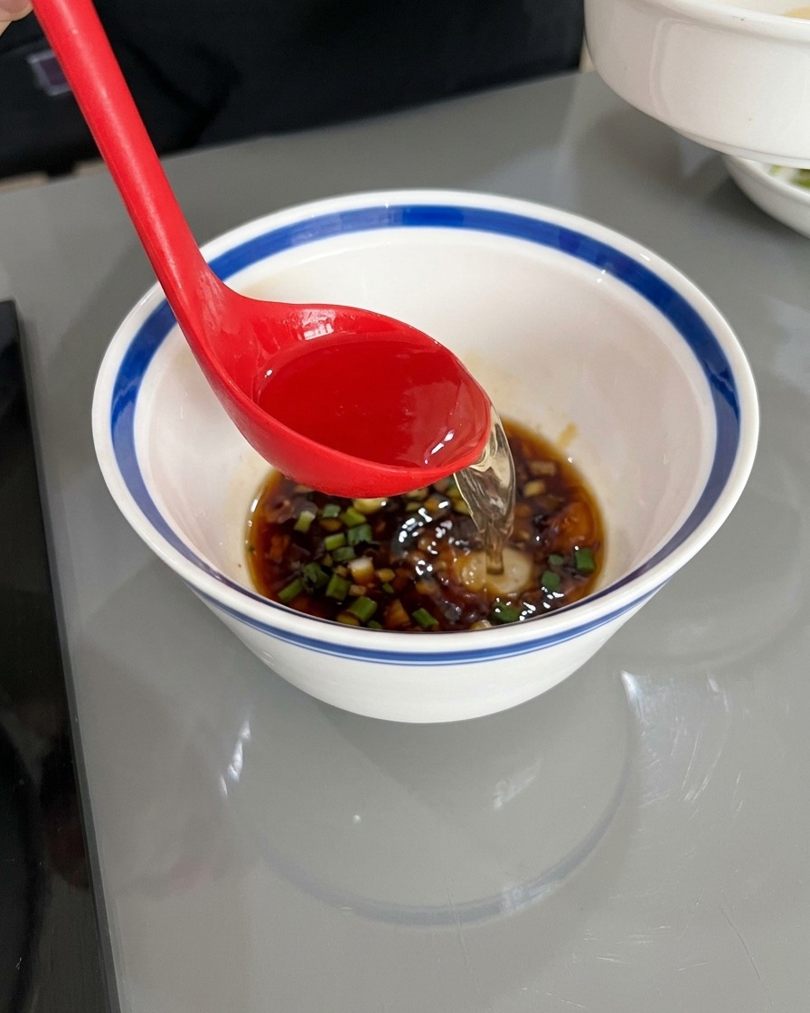 A red ladle pouring clear hot water into a white bowl containing dark soy sauce, oil, and chopped aromatics.