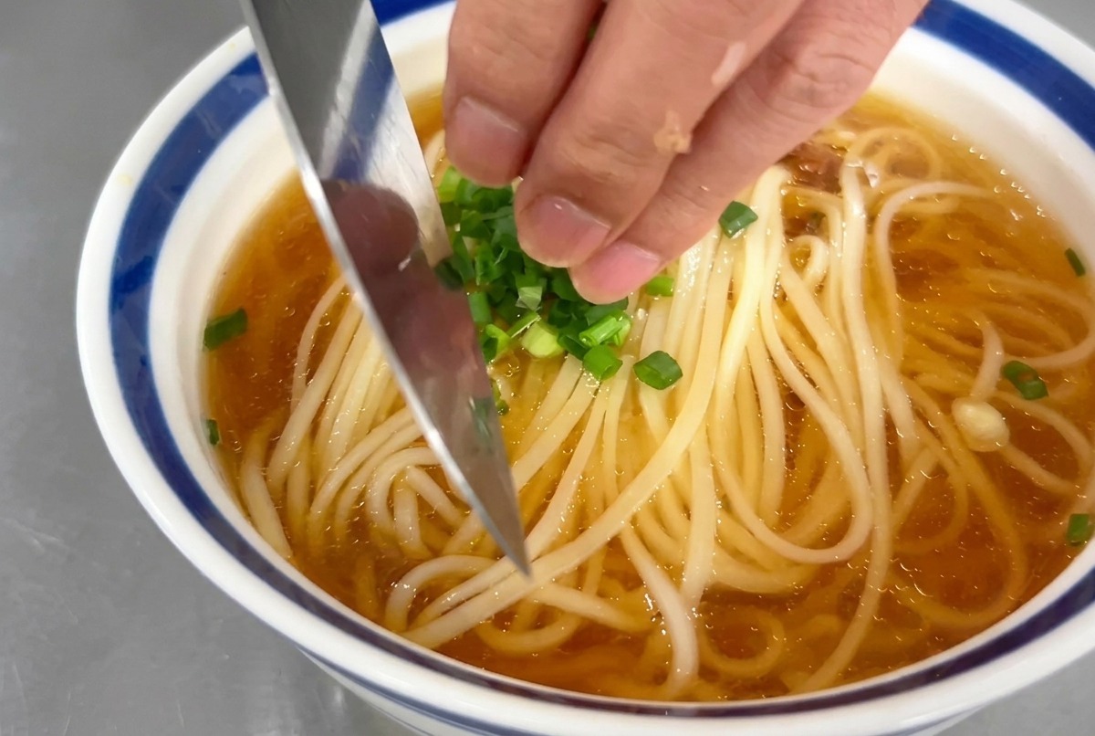 Hand sprinkling small rings of green scallions onto a bowl of noodle soup.