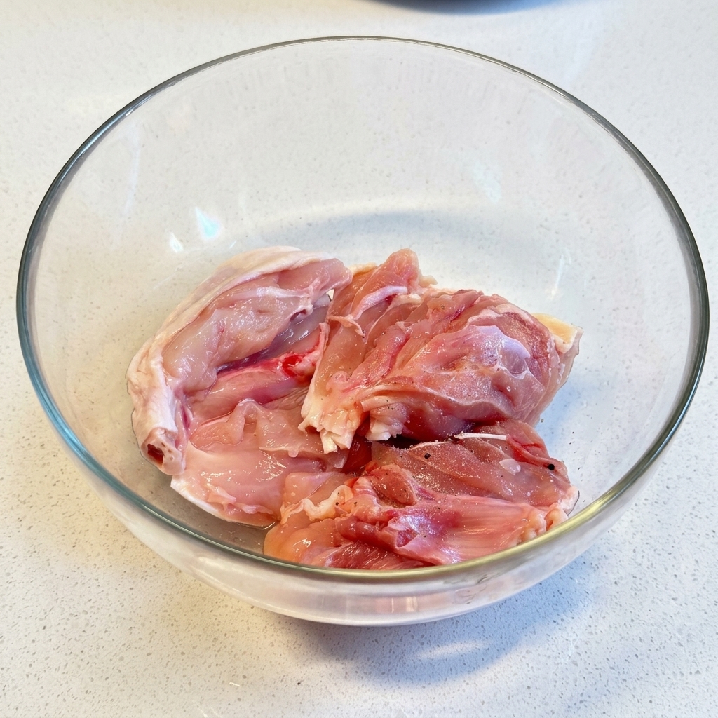 Raw chicken thighs being sprinkled with salt and pepper inside a clear glass mixing bowl.