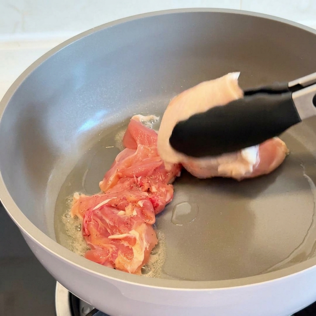 Tongs placing a raw chicken thigh skin-side down into a grey non-stick pan with shimmering oil.
