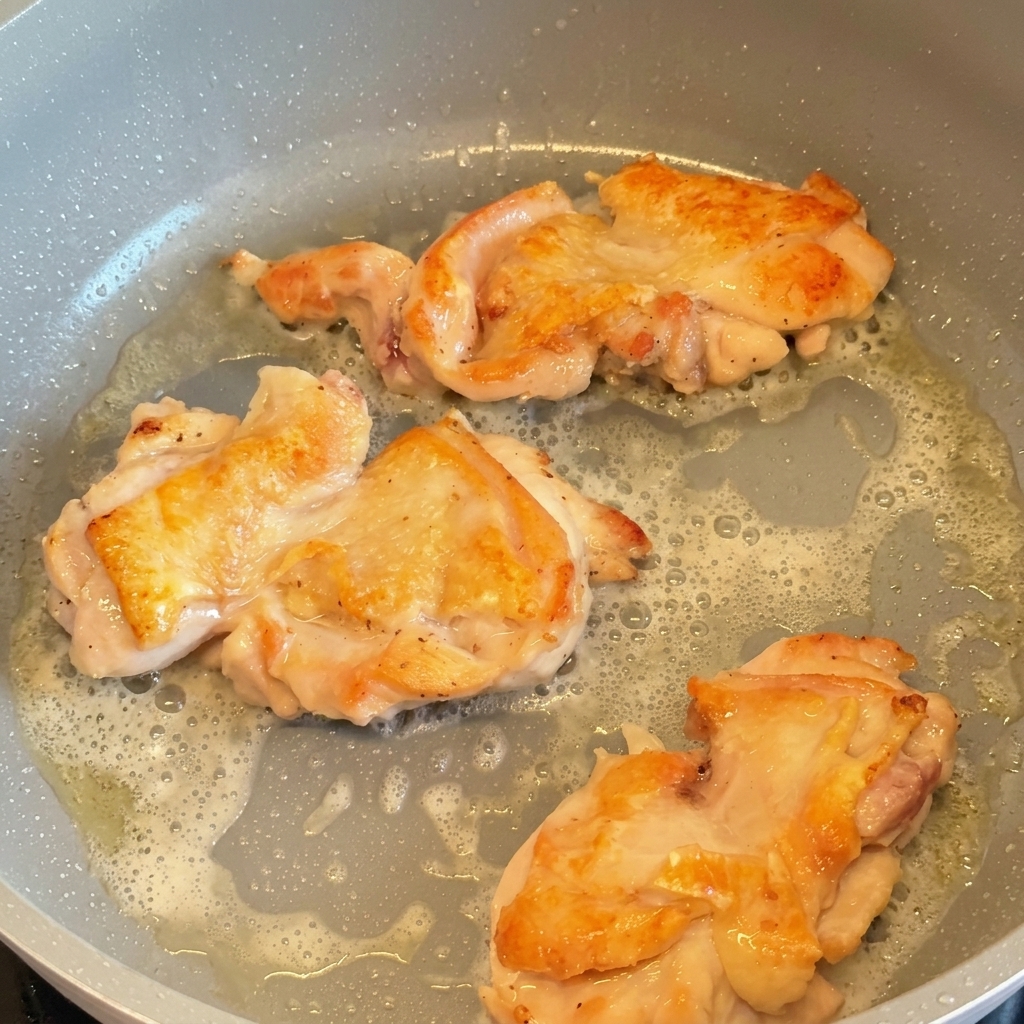 Three chicken thighs frying in a pan, flipped over to show their crispy, golden-brown seared skin.