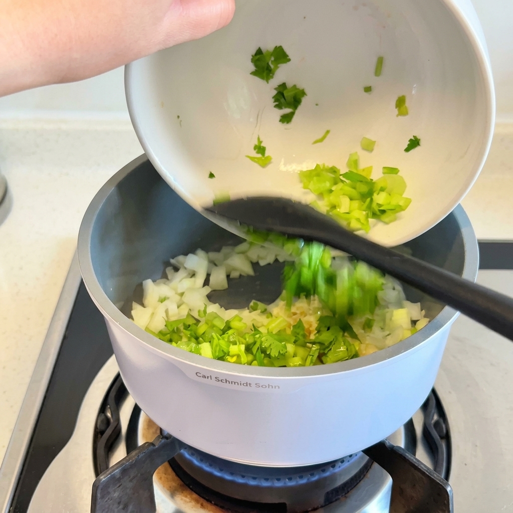 Chopped celery and fresh herbs being poured from a white bowl into a cooking pot containing translucent onions.