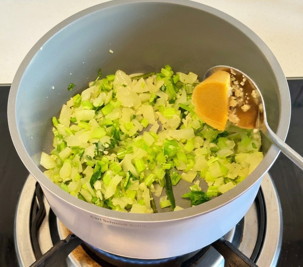 A metal spoon adding a yellow concentrated chicken bouillon cube into a grey pot with sautéed onion and celery pieces.