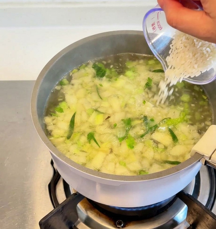 White rice being poured from a clear measuring cup into a pot of simmering soup with visible chopped vegetables and herbs.