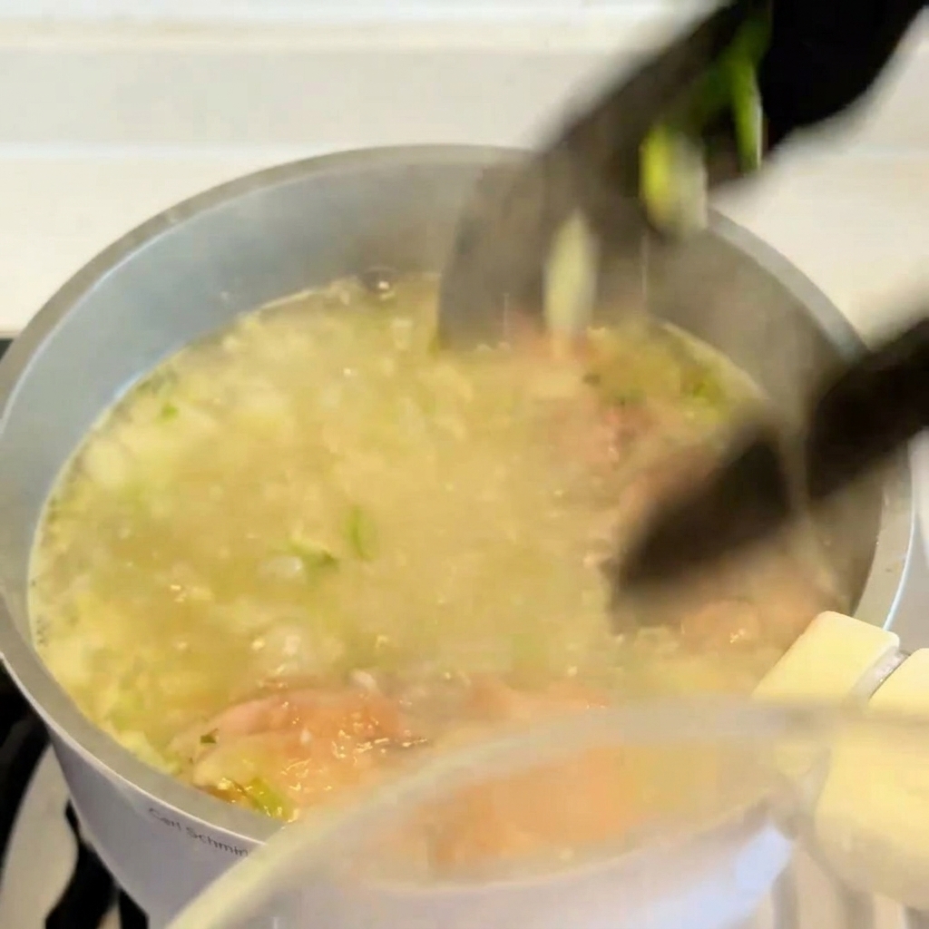 A pair of tongs lifting a cooked, golden-brown chicken thigh out of a pot of simmering soup broth.