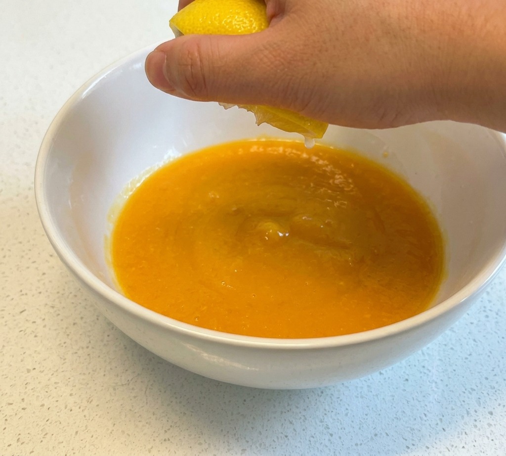 A hand squeezing juice from a yellow lemon half into a white ceramic bowl containing beaten eggs.