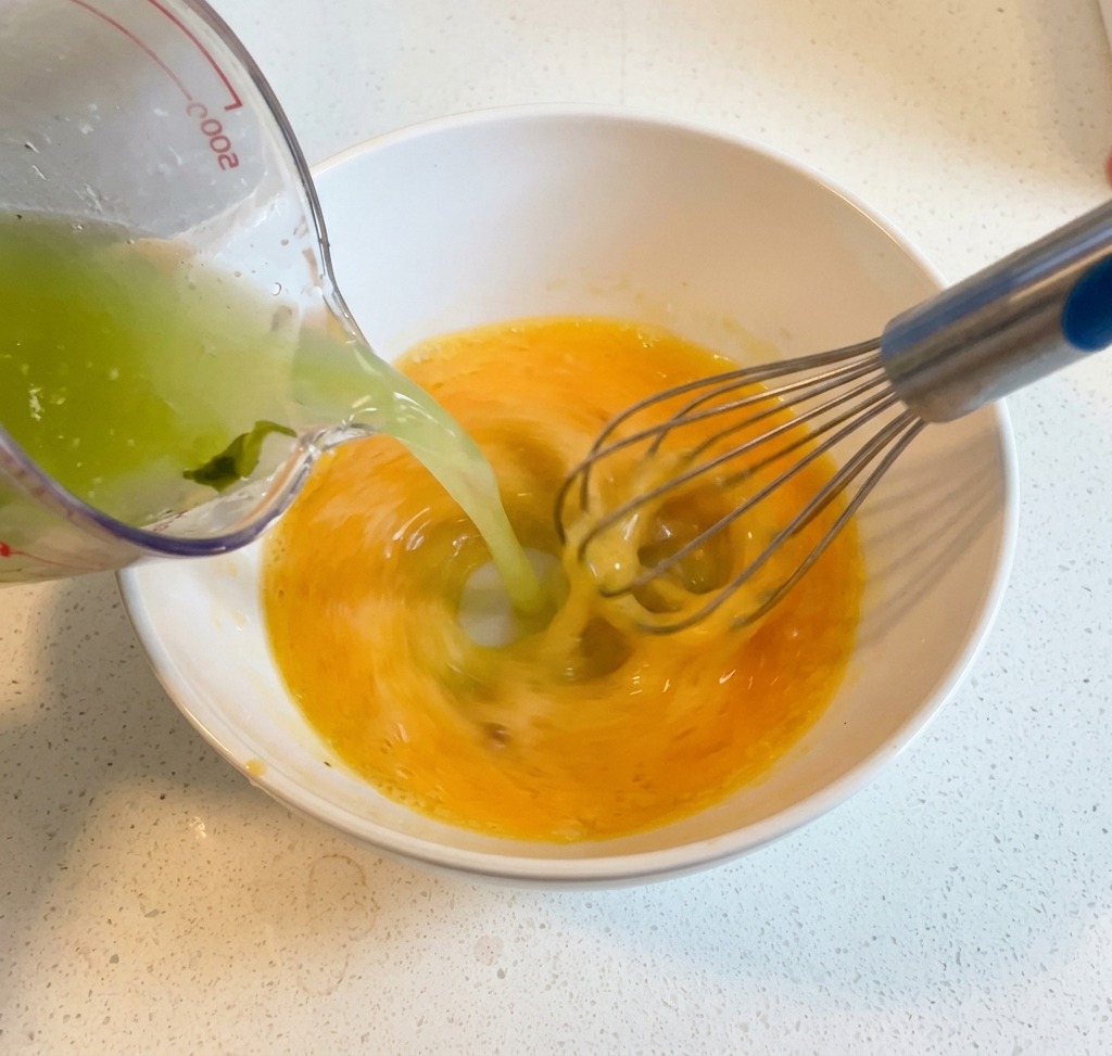 Hot broth being poured from a measuring cup into a bowl of whisked eggs and lemon while being mixed with a wire whisk.