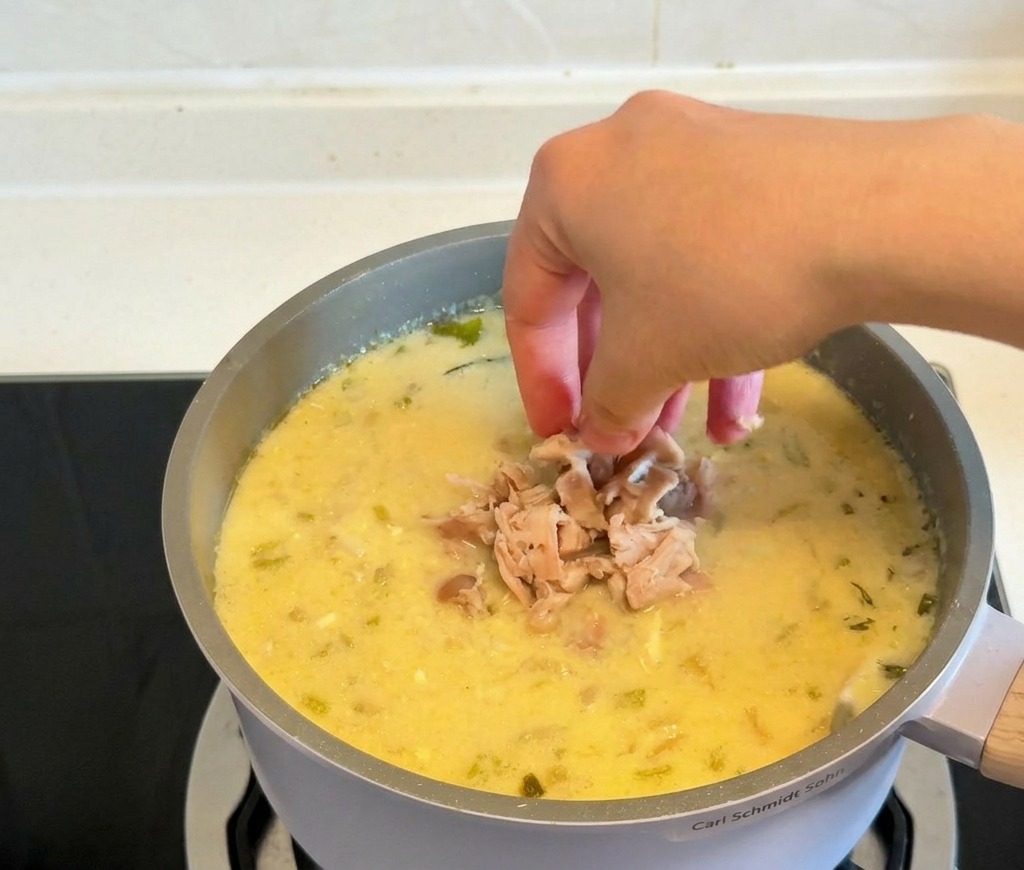 Hands dropping piles of shredded cooked chicken into a pot of thickened creamy yellow soup.