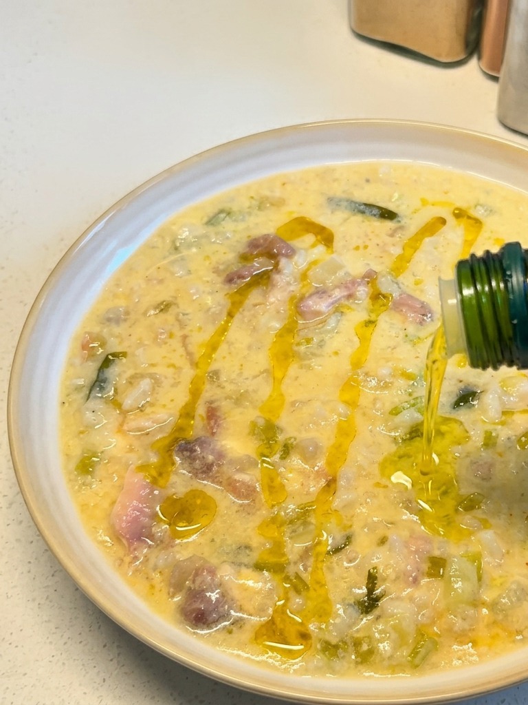Golden olive oil being drizzled from a green bottle onto a plated bowl of creamy lemon chicken soup.