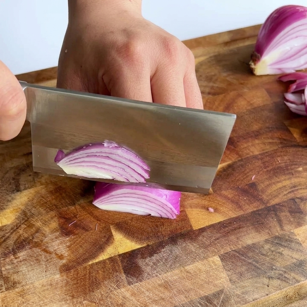 A large cleaver slicing through a red onion creating thin purple slivers on a wooden cutting board.
