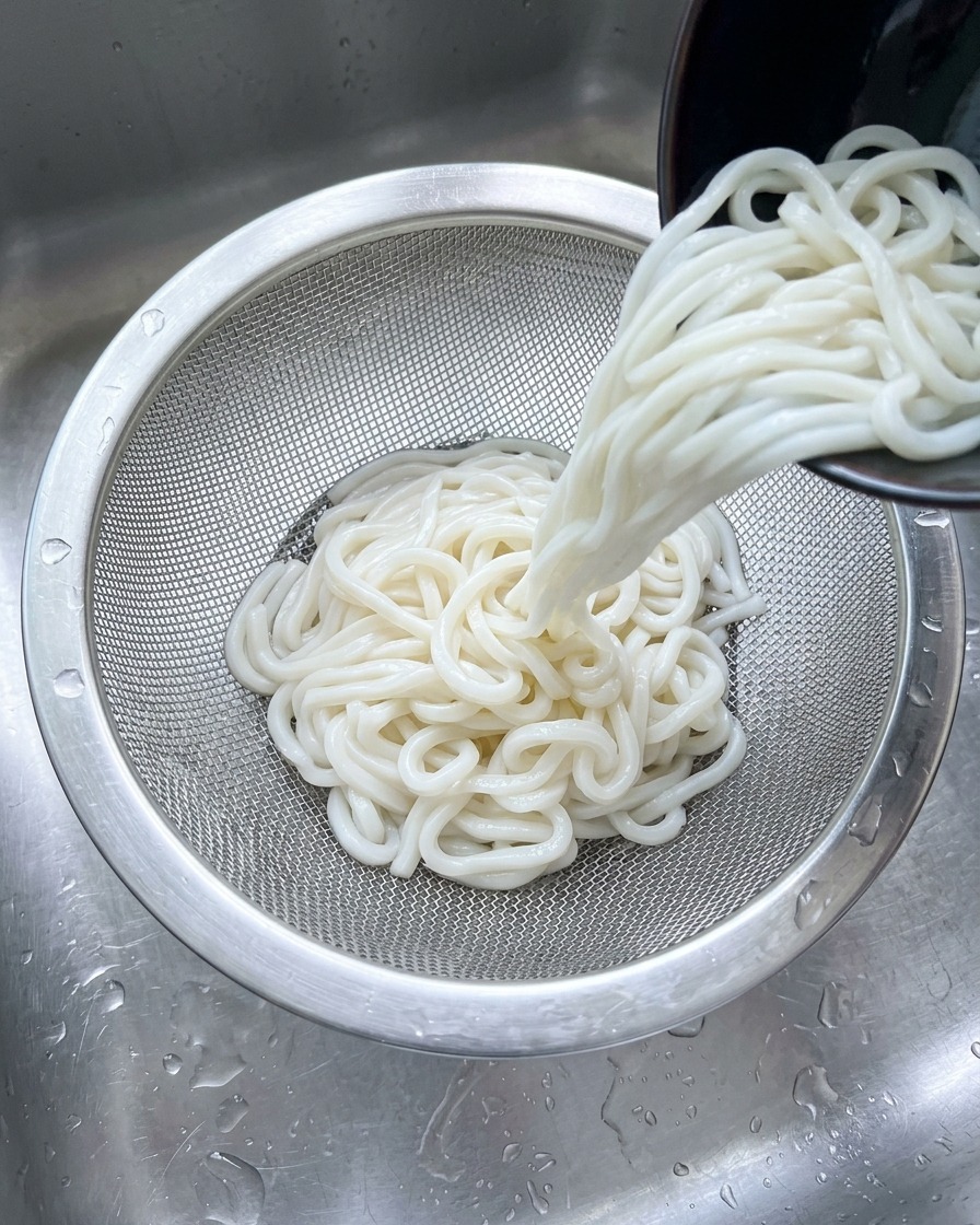 White udon noodles being poured into a stainless steel mesh strainer under a stream of cold water in a sink.