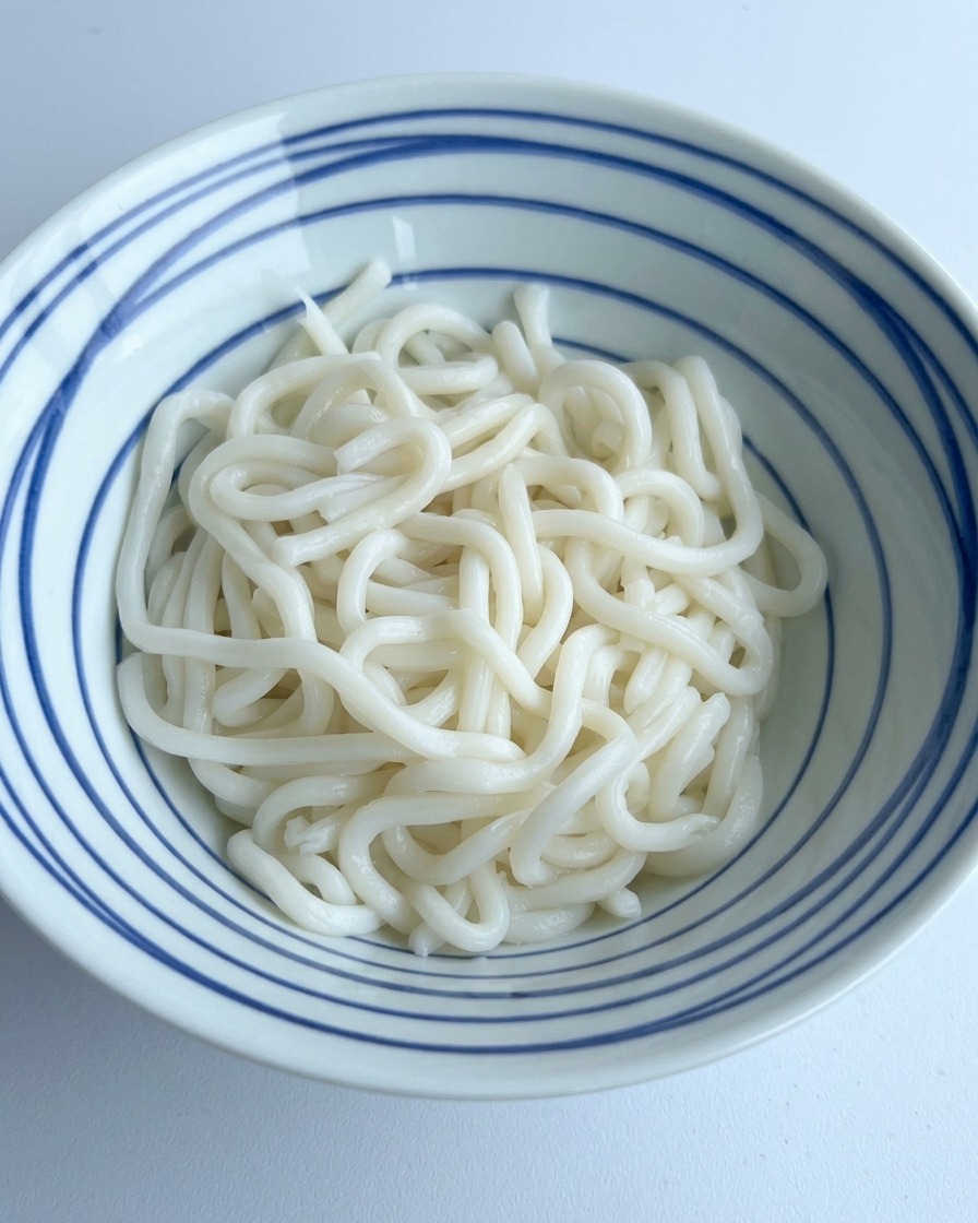 Dark soy sauce and seasonings being poured over a pile of white udon noodles in a blue-striped bowl.