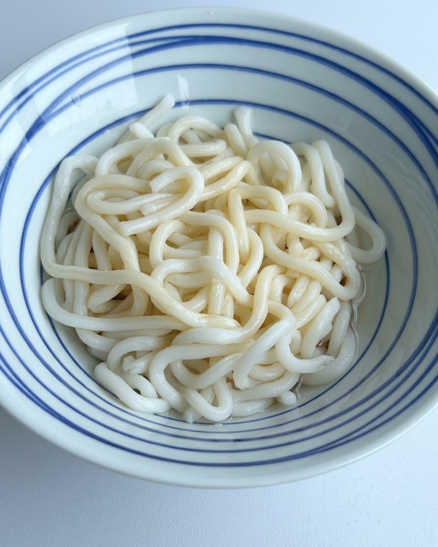 Clear water being poured into a bowl of udon noodles, mixing with the dark seasonings at the bottom.